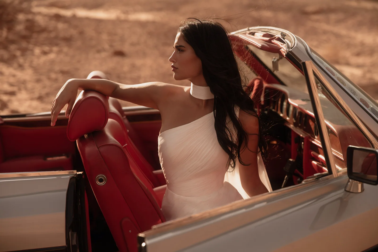Bride wearing a designer wedding dress beside a red-interior convertible